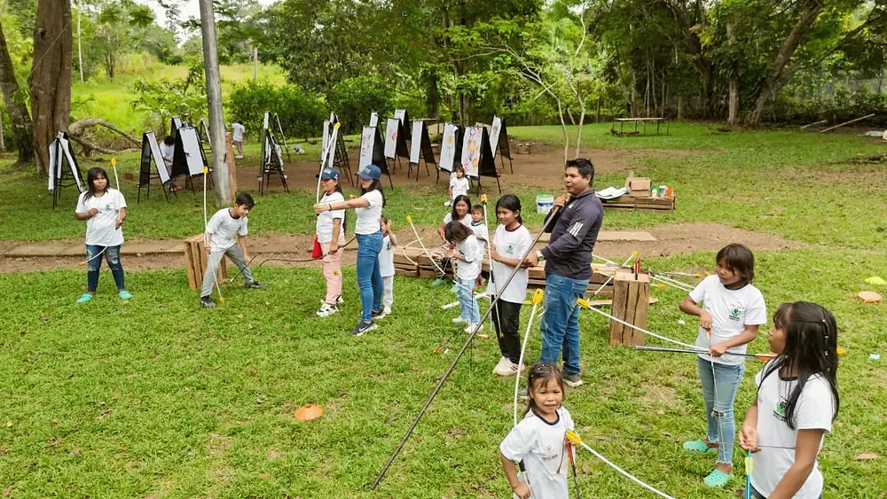 Niñas y niños con arcos en un prado, con dianas colgadas entre los árboles.