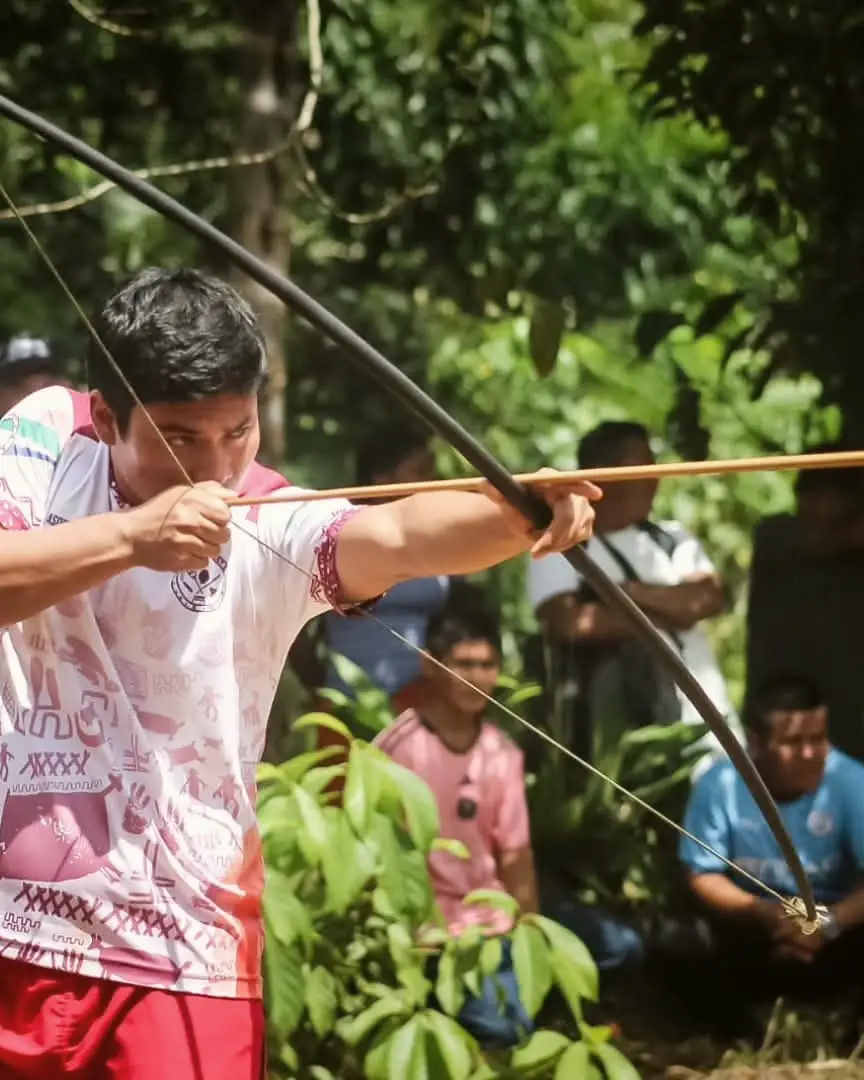 Un joven tensa un arco largo en medio de la selva; detrás, otras personas observan.